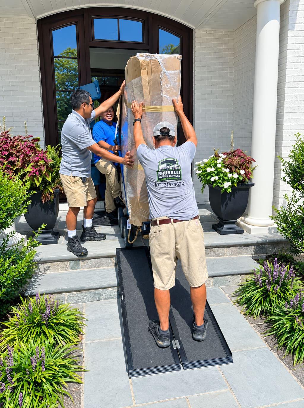 Two Brunalli movers carrying a tall wrapped piece down a ramp at a front door